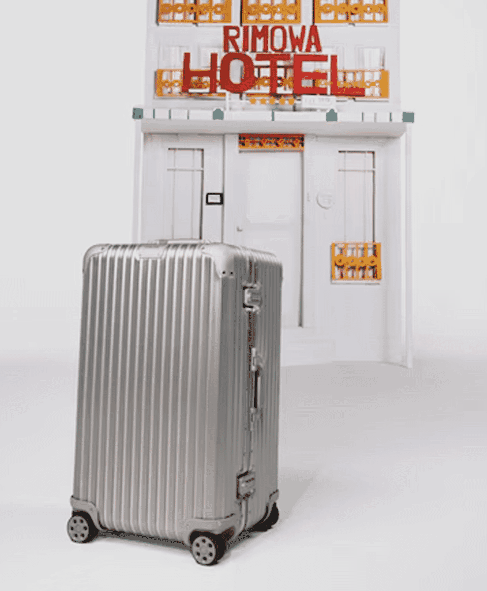 A silver Rimowa suitcase standing next to a miniature white hotel model representing high-end hospitality.