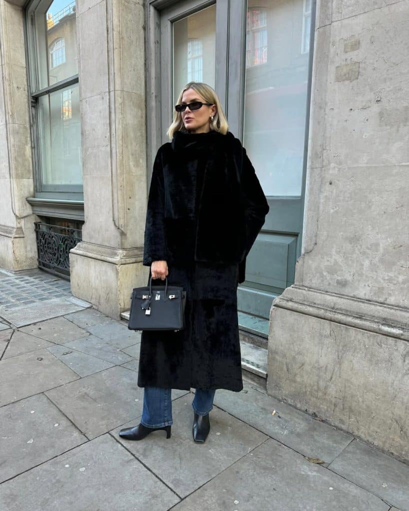A woman in a black faux fur coat and blue jeans standing on a city sidewalk wearing black ankle boots.