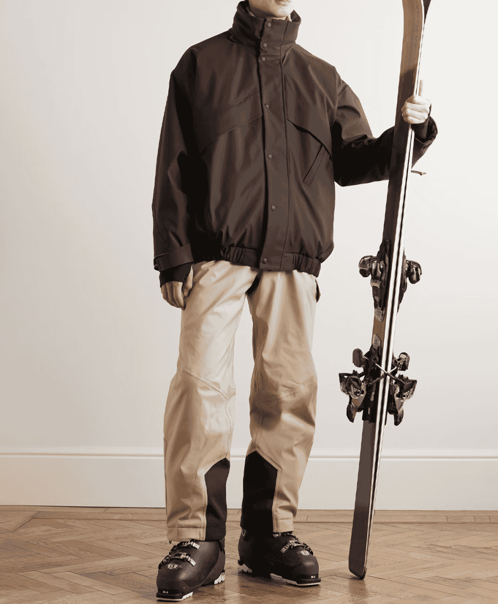A model in a brown technical jacket and beige pants holding skis indoors.