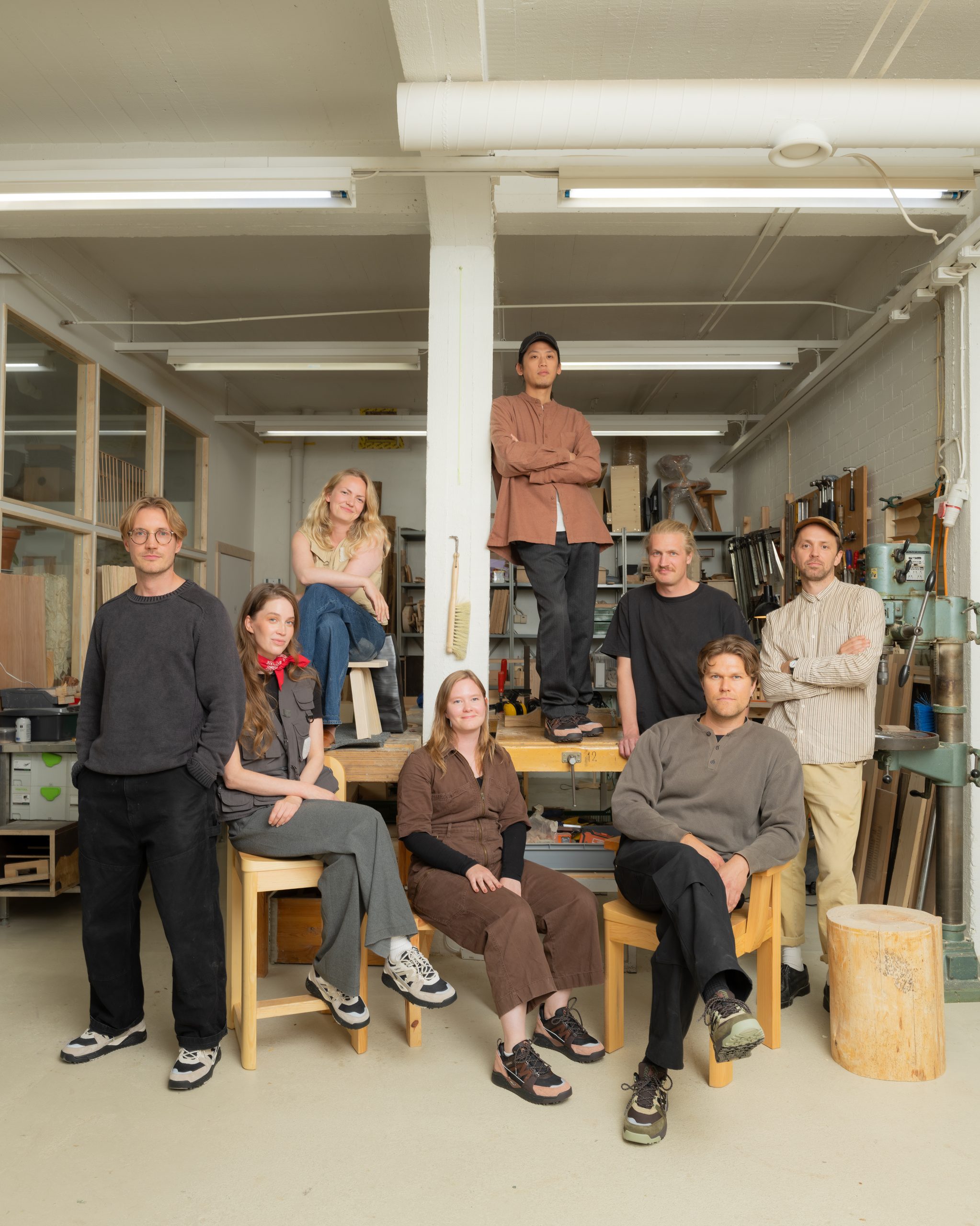 A group of seven people in a woodworking workshop wearing earth-toned casual and utility clothing.
