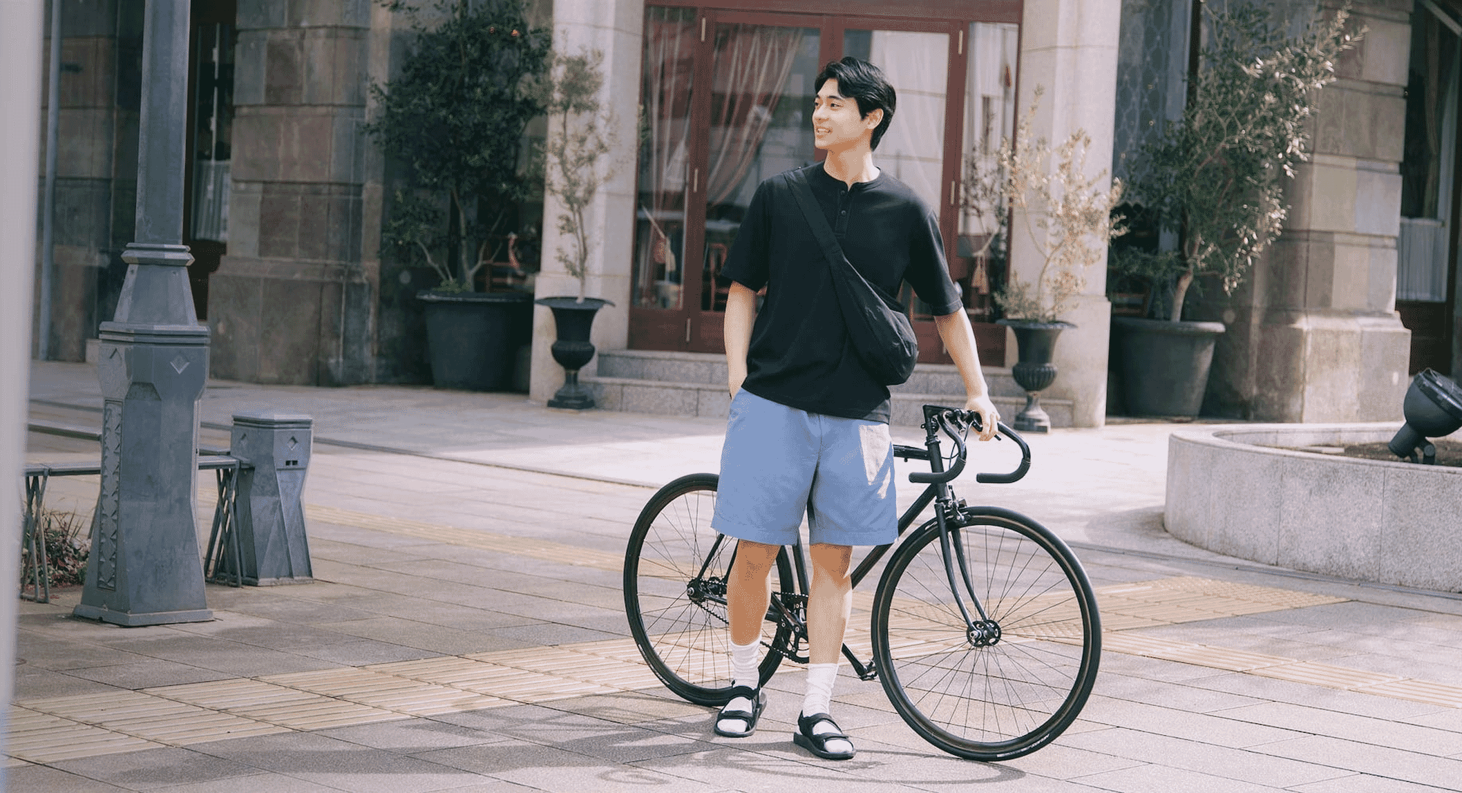 Full-body shot of a man in a black henley and light blue shorts standing with a bicycle on a city sidewalk.