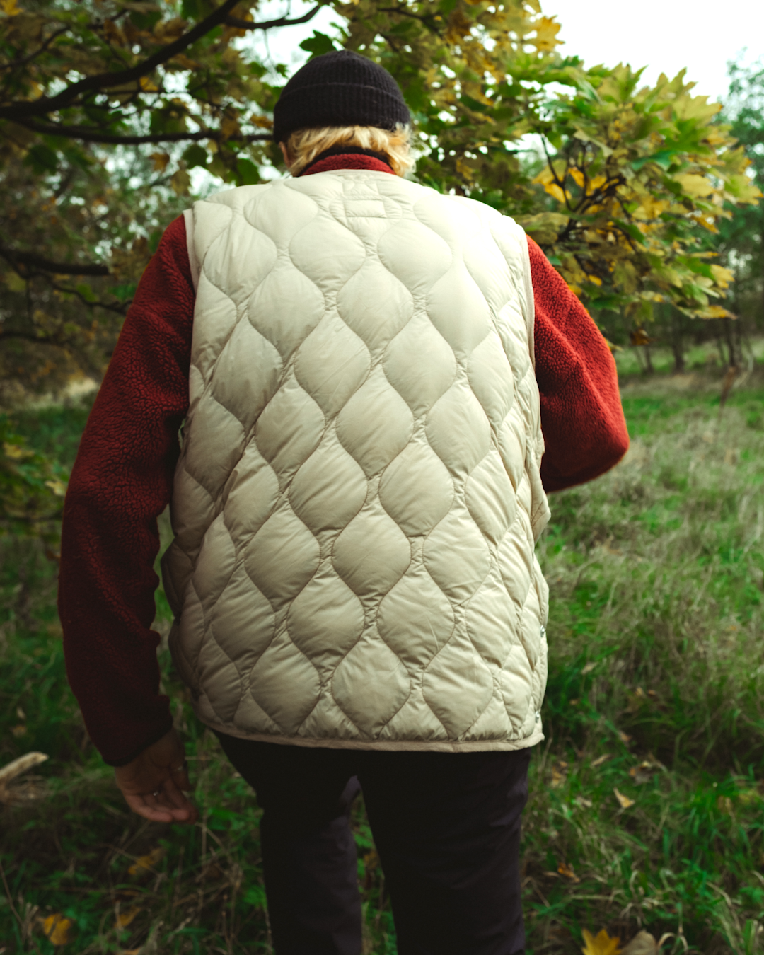 Rear view of a person in a cream diamond-quilted vest over a red fleece in a forest.
