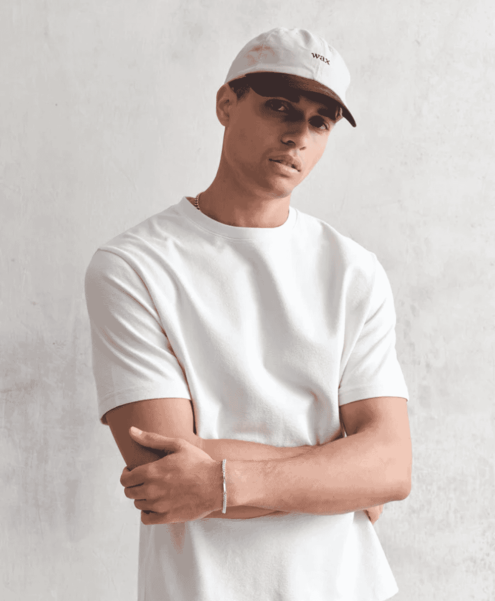 Man in a white t-shirt and white baseball cap with 'wax' embroidery posing against a white wall.
