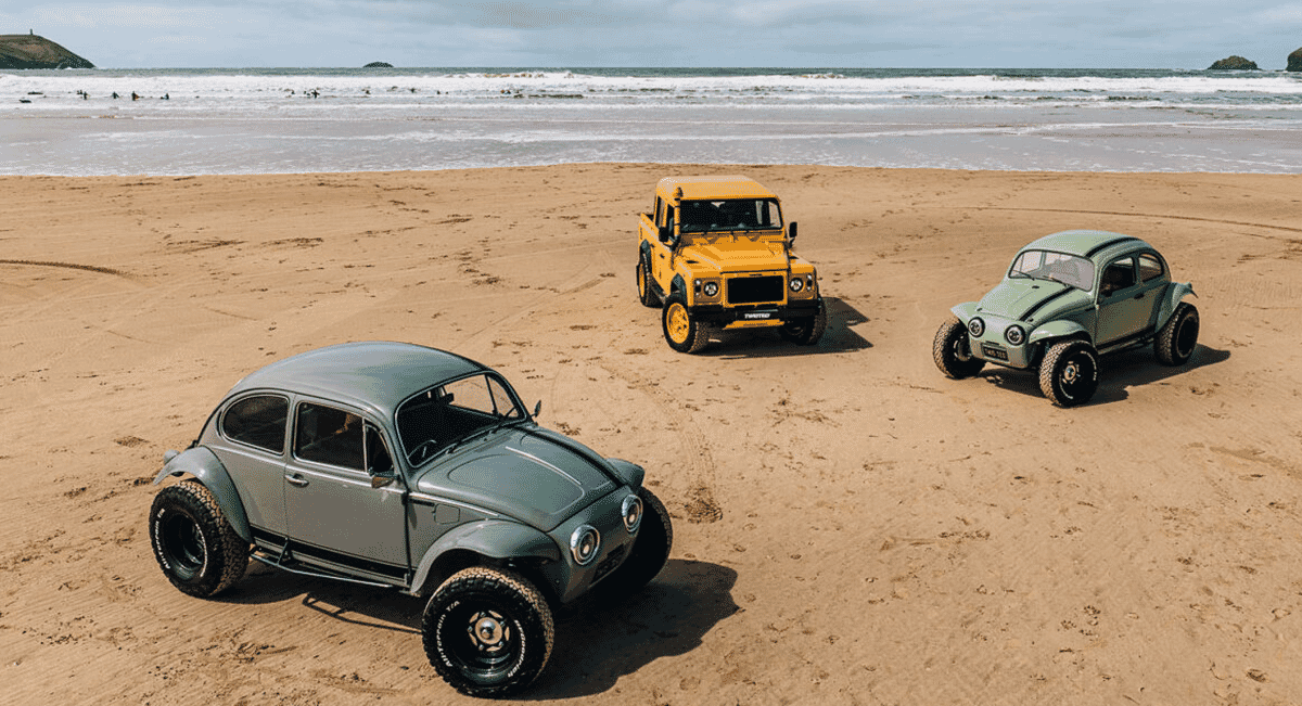 Three customized vehicles, including two Beetles and a Land Rover, parked on a sandy beach near the surf.