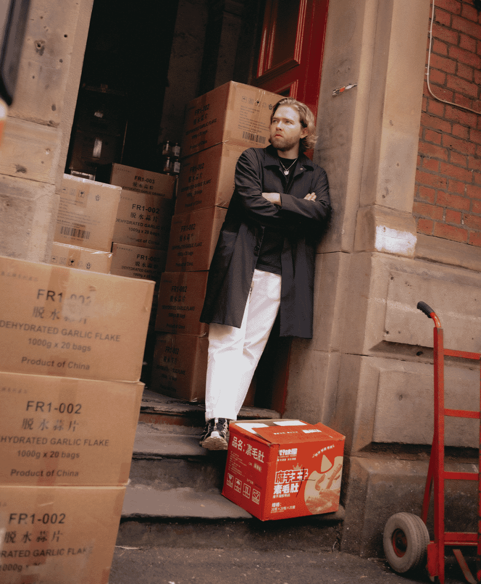 Full-body shot of a man wearing a black coat and white wide-leg pants in an urban alley.