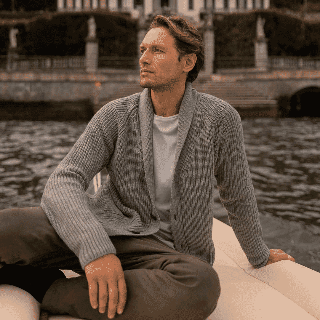 A man in a grey ribbed cardigan on a boat with a lakeside villa background.