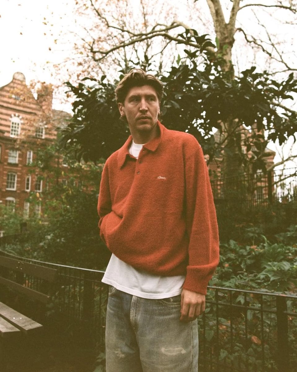 Man in a textured orange knit polo sweater in an autumnal park.