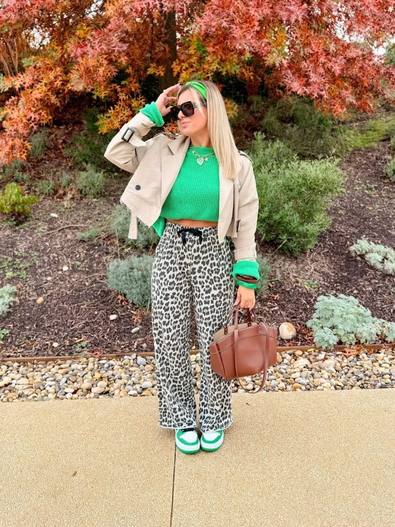 Woman in a green cropped sweater and leopard pants posing against autumn foliage.