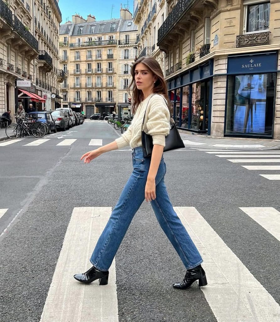 A woman crossing a Parisian street in a cream knit sweater, high-waisted blue jeans, and black patent leather Chelsea boots.