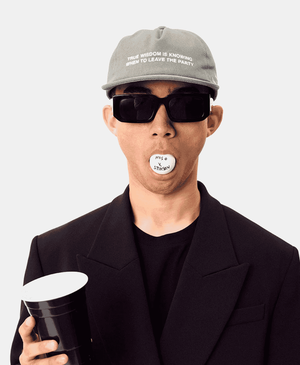 Man wearing a gray Stiksen slogan cap, black sunglasses, and a structured black blazer while holding a branded candy.