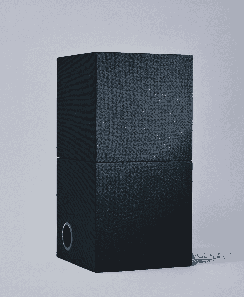 Two black fabric-covered speakers stacked vertically in a minimalist studio setting.