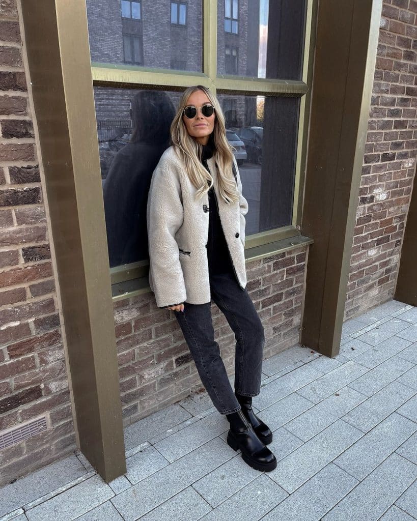 A woman in a beige shearling coat, dark jeans, and chunky black boots leaning against a brick wall.