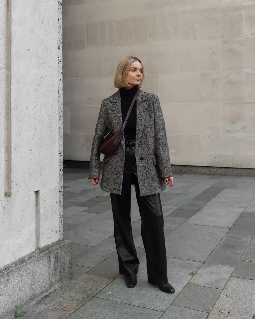 A woman in a grey textured blazer, black leather pants, and a burgundy bag standing on an urban sidewalk.