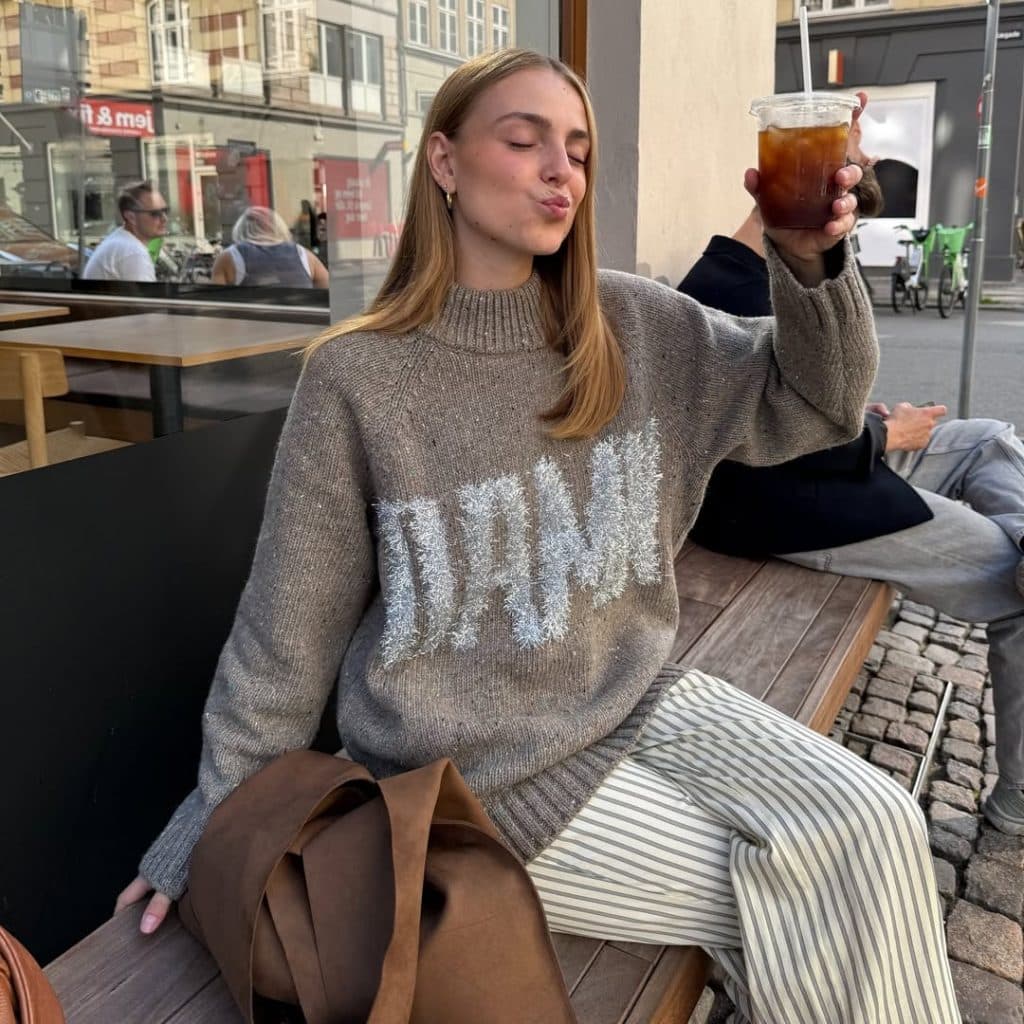 A woman in a grey 'DAMN' knit sweater and striped pants sitting at a café.
