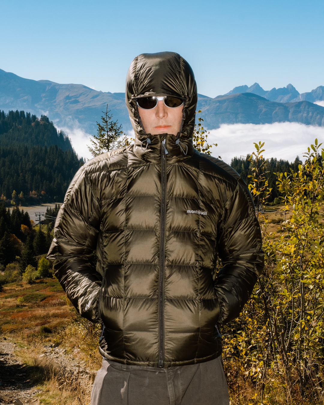 A man in a dark olive Montbell puffer jacket standing against a mountain vista with clouds.