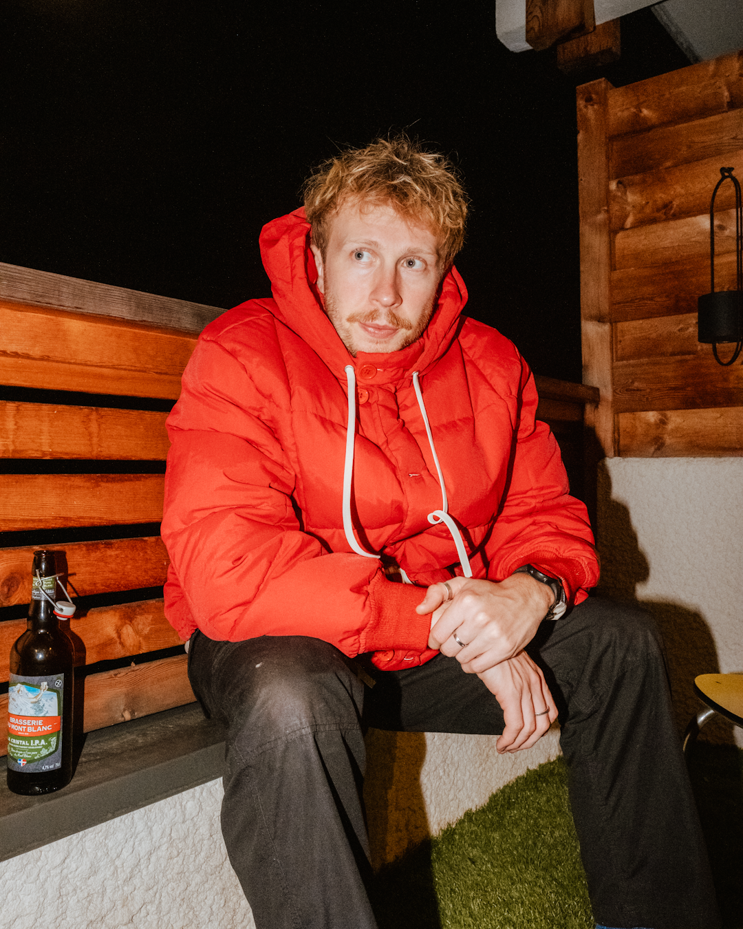 A man in a red hooded puffer jacket sitting on a wooden bench at night with a beer bottle.