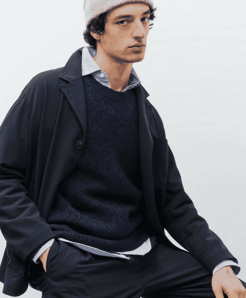 A man in a dark navy knit sweater, black coat, and a beige beanie in a studio setting.
