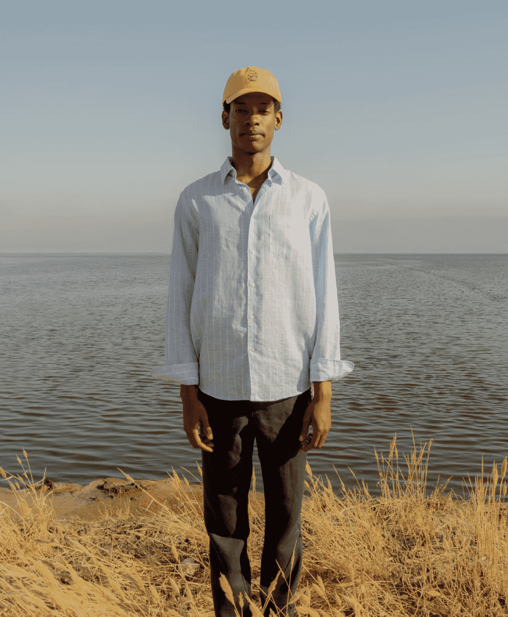 Man by the water wearing a mustard yellow cap and a striped blue shirt.