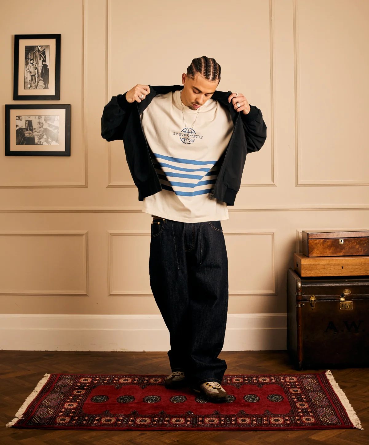Model wearing a black bomber jacket over a striped graphic tee with baggy dark jeans and patterned sneakers.