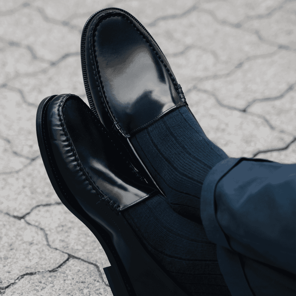 Close-up of black glossy loafers and navy trousers on stone pavement.