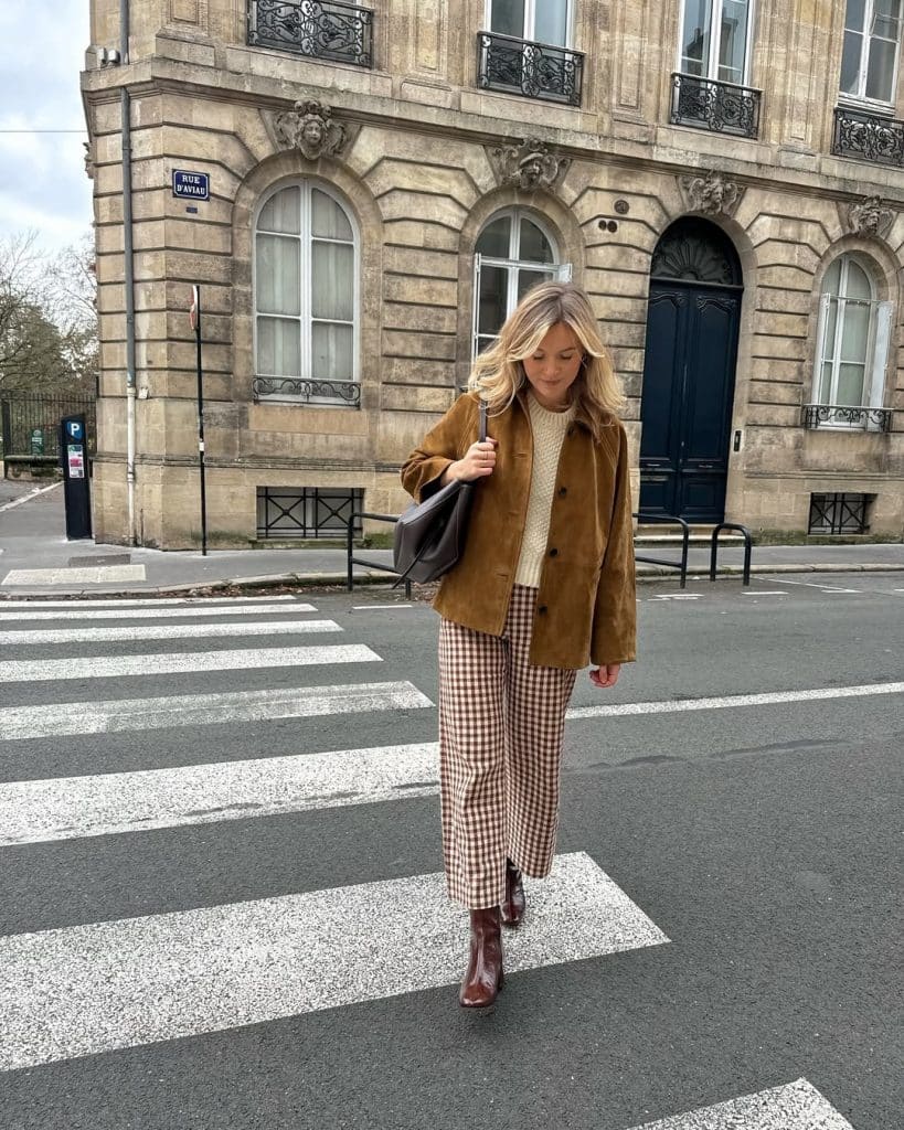 A woman in a brown suede jacket, gingham trousers, and brown boots crossing a street in Paris.