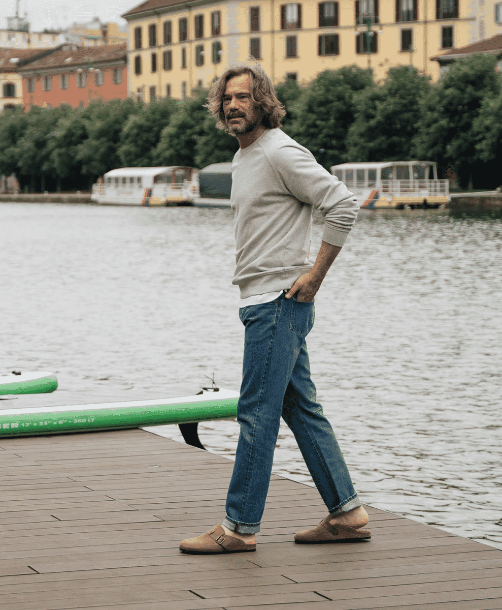 A man in denim and clogs walking on a wooden dock by a river.