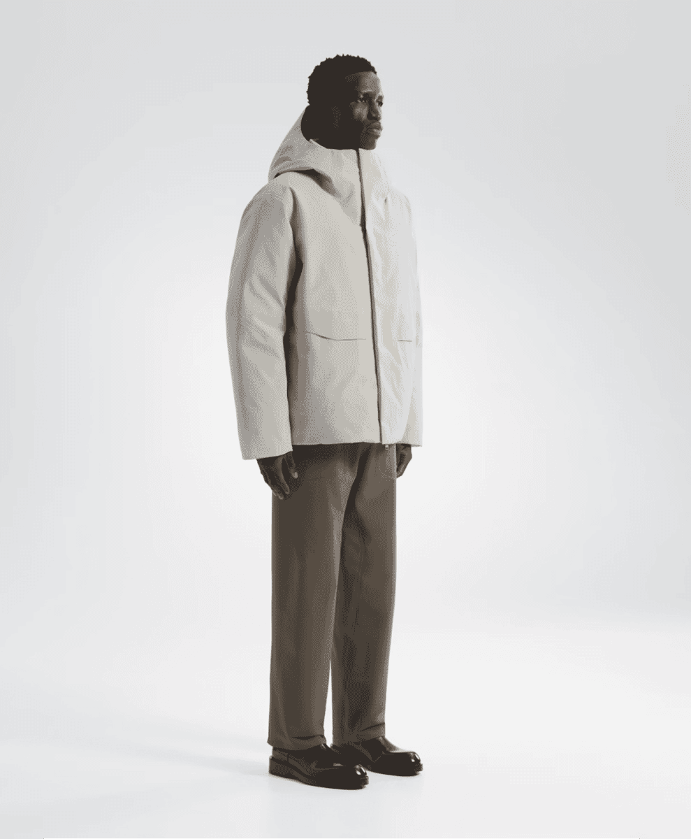 A man in a minimalist white hooded puffer coat and dark trousers standing in profile against a white background.