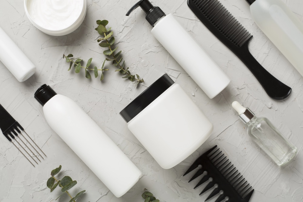 A flat-lay of white beauty containers, black combs, and eucalyptus on a textured gray background.