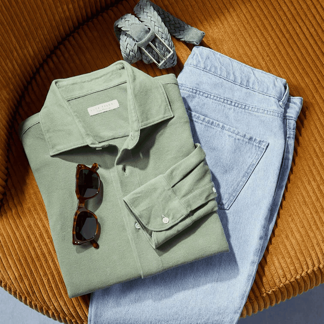 Folded sage green button-up shirt and denim jeans on a corduroy chair.