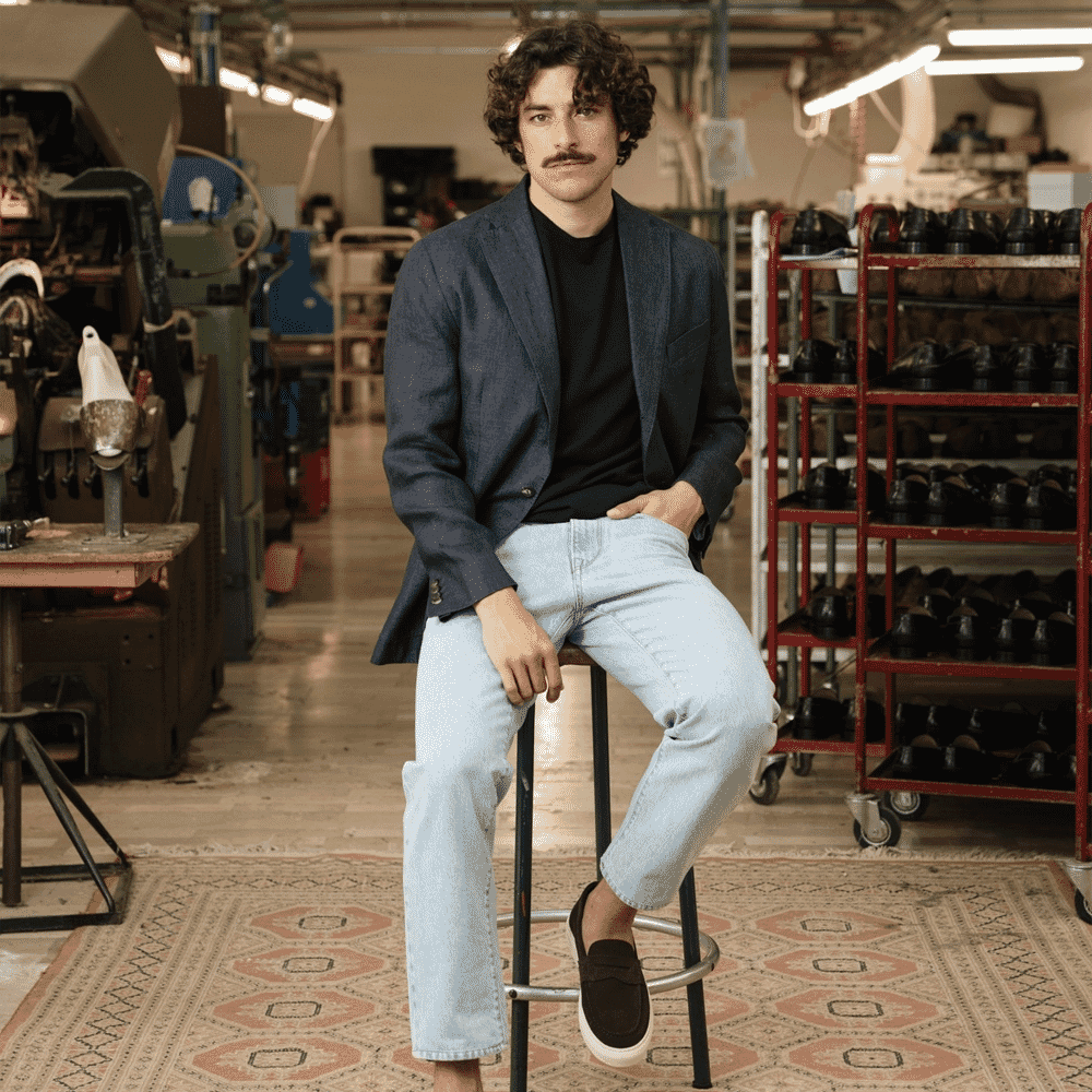 A man in a navy blazer and jeans in a shoemaker's workshop.