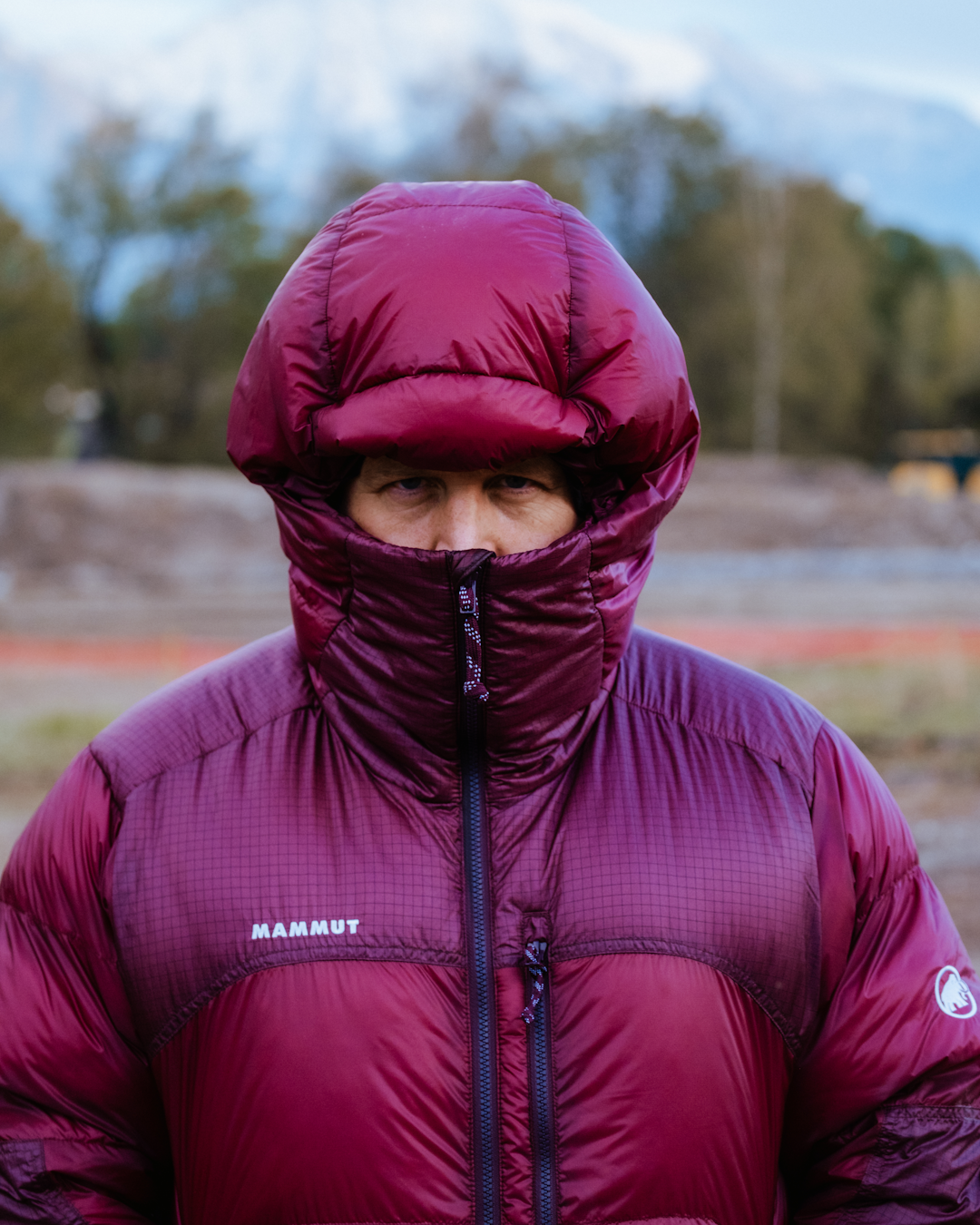 Close-up of a maroon Mammut puffer jacket with the hood up against a blurred mountain background.