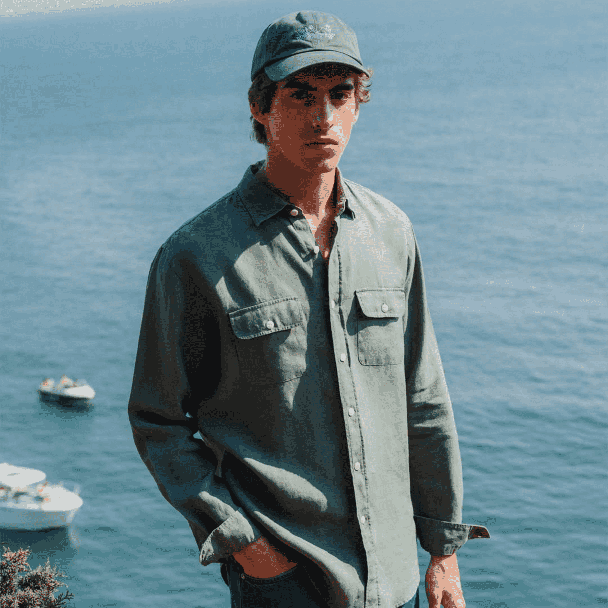 A man in an olive green button-up shirt and cap by the sea.