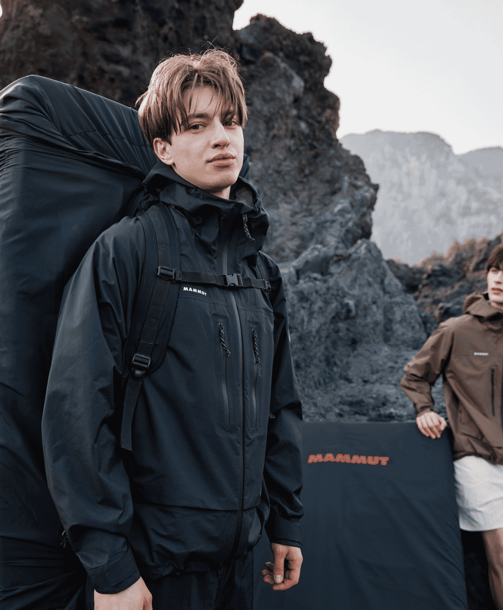 A man in a black Mammut technical jacket against a rugged mountain landscape.