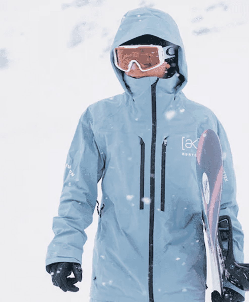 A snowboarder in a light blue hooded jacket holds a snowboard in falling snow.