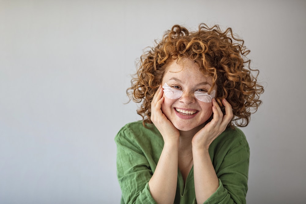 A woman with curly red hair wearing skincare eye patches and a green linen-style top.