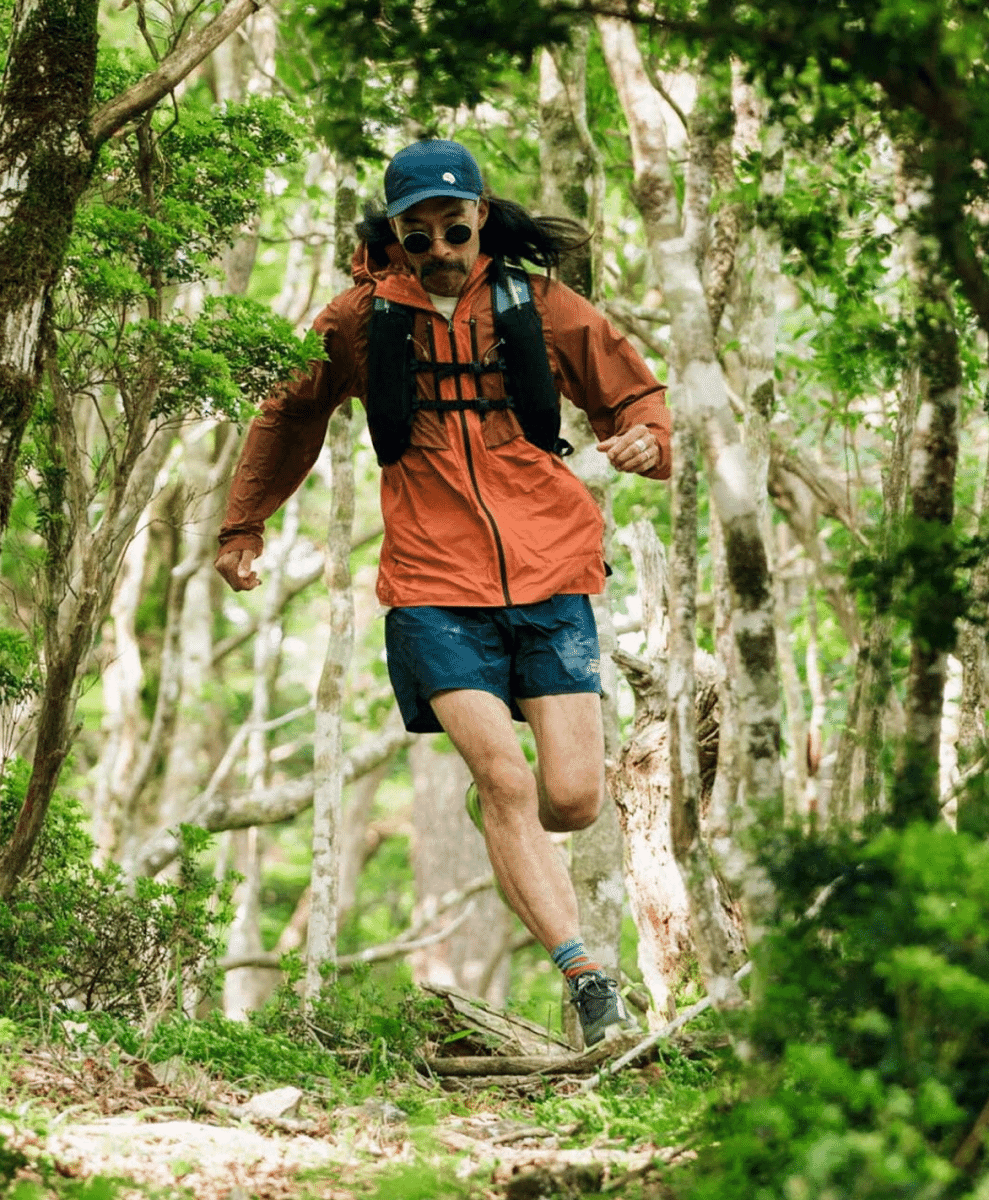 A male runner in a bright orange rain jacket and hydration pack sprinting through a forest trail.