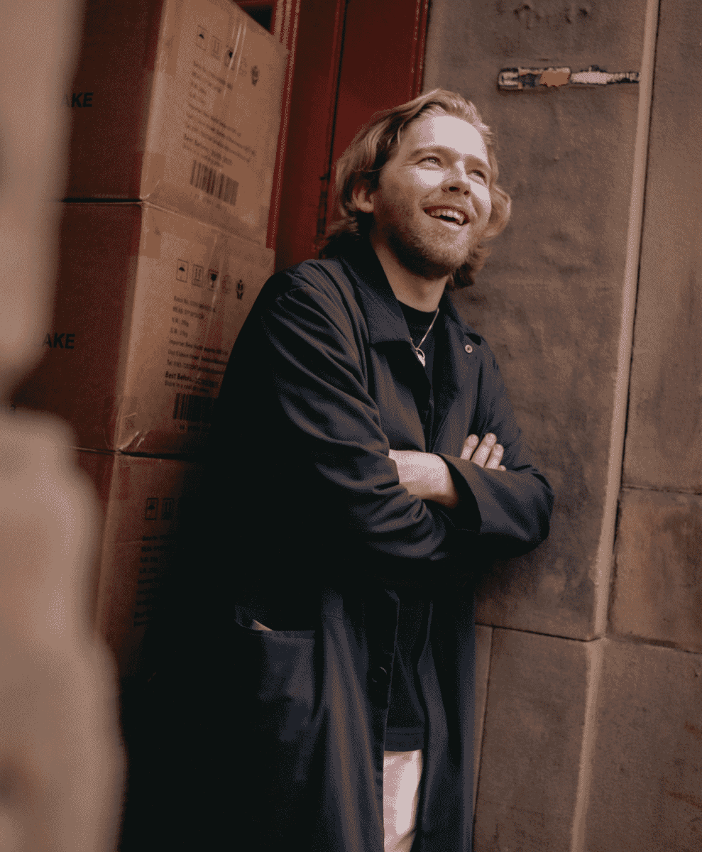 Man in a dark trench coat leaning against a brick wall with cardboard boxes.