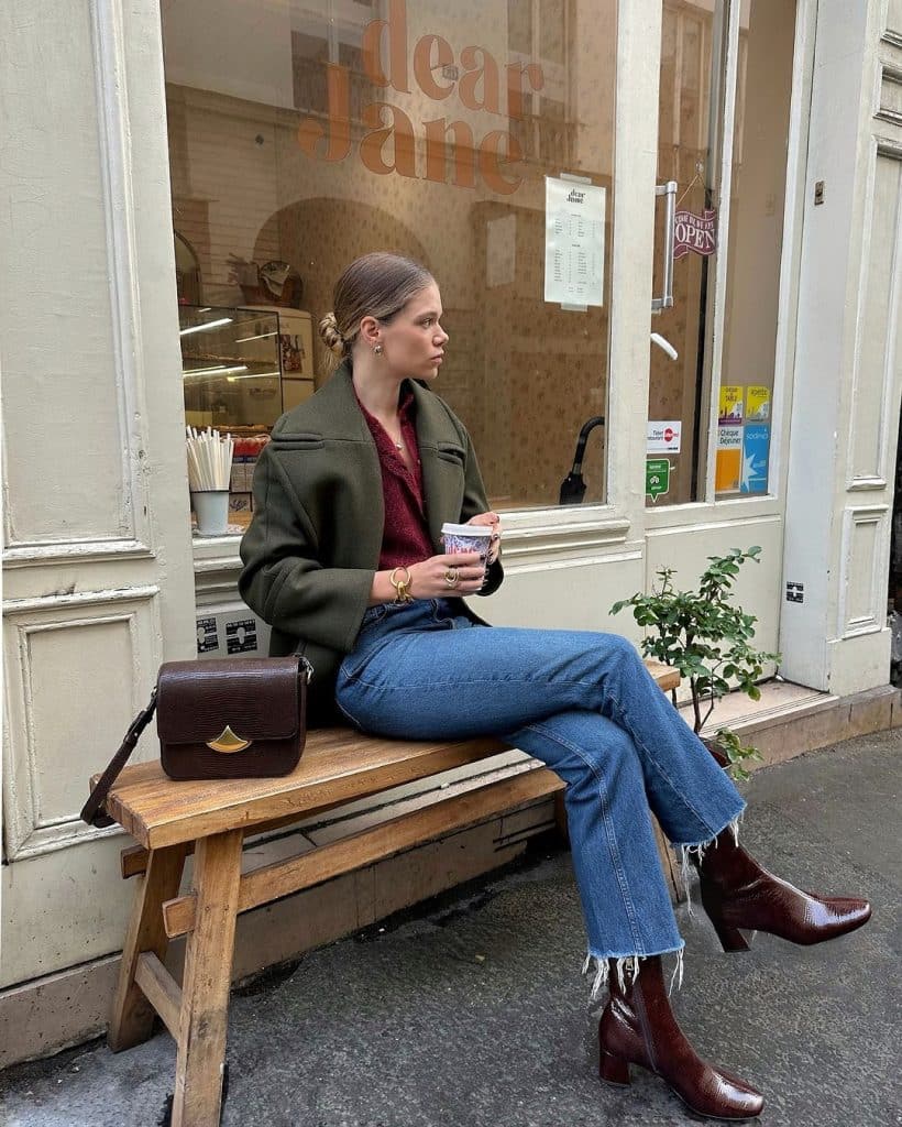 A woman in an olive green coat and red top with flared blue jeans and brown boots sitting outside a cafe.