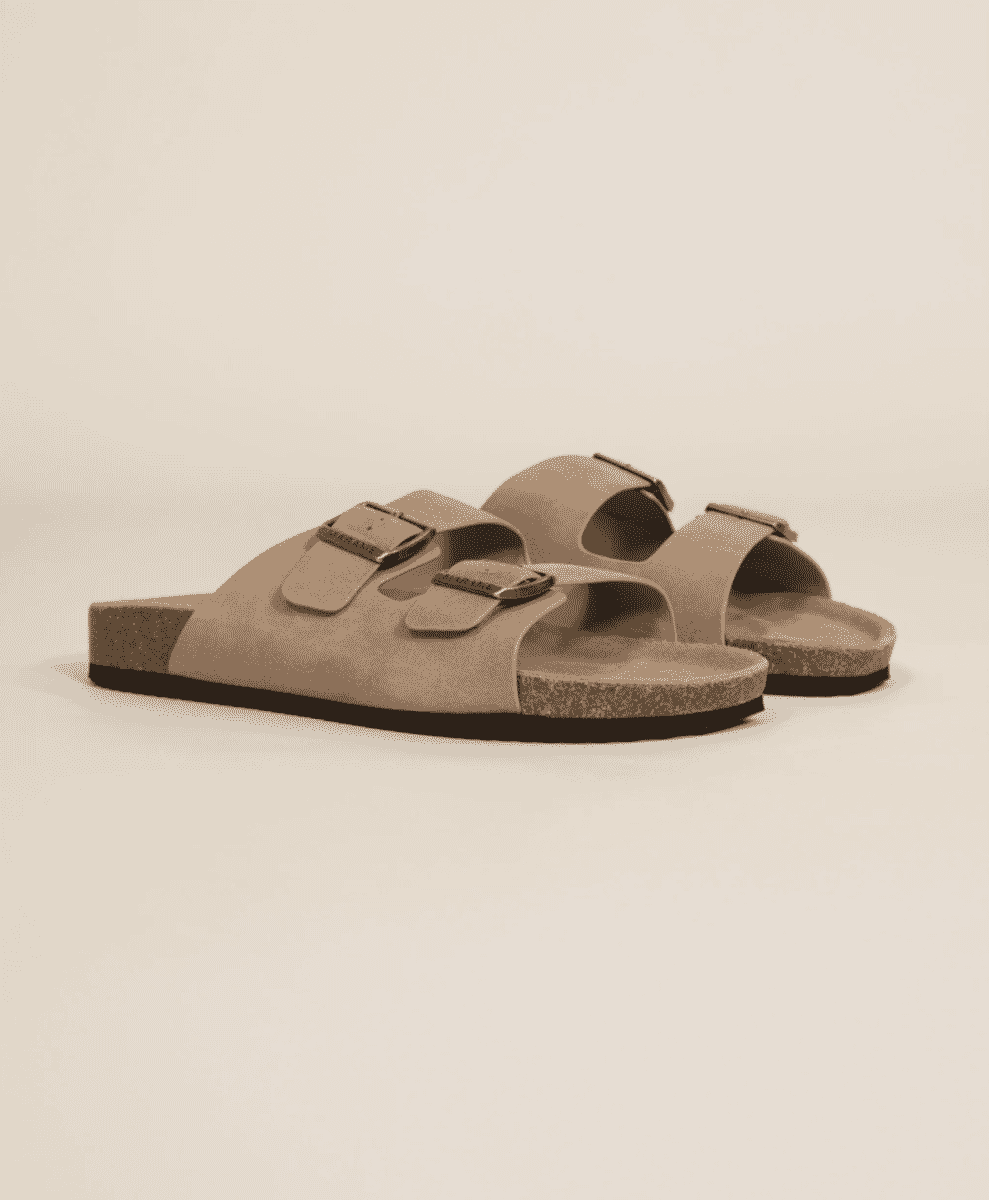 A pair of beige suede double-strap sandals with metal buckles on a neutral background.
