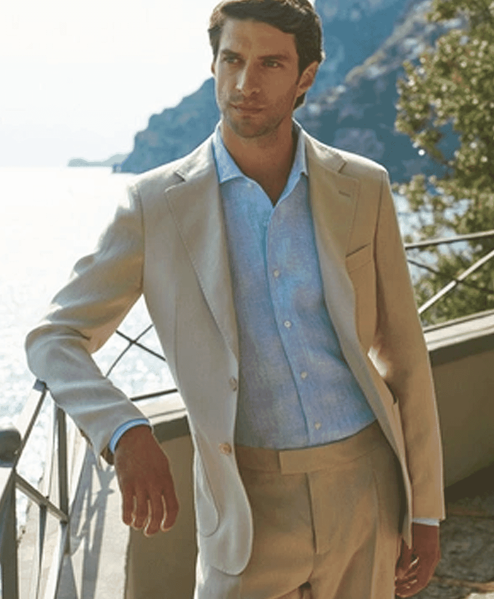 A man in a light beige linen blazer on a sunlit balcony overlooking a coastal landscape.