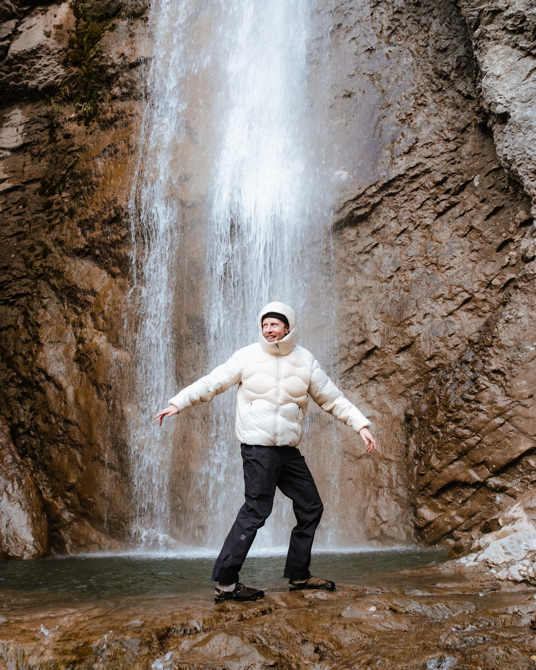 A man in a white puffer jacket joyfully standing under a waterfall with arms outstretched.