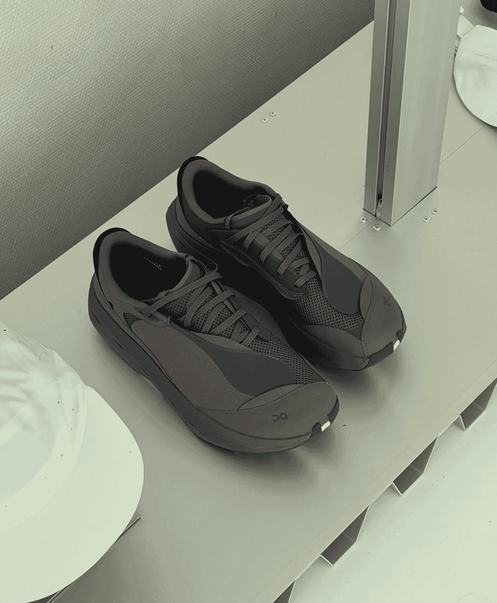 Dark gray On running shoes displayed on a minimalist white shelf alongside white caps.