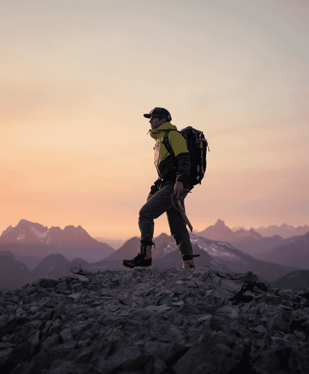 A hiker in a green and black technical jacket walking across a mountain ridge at sunset.