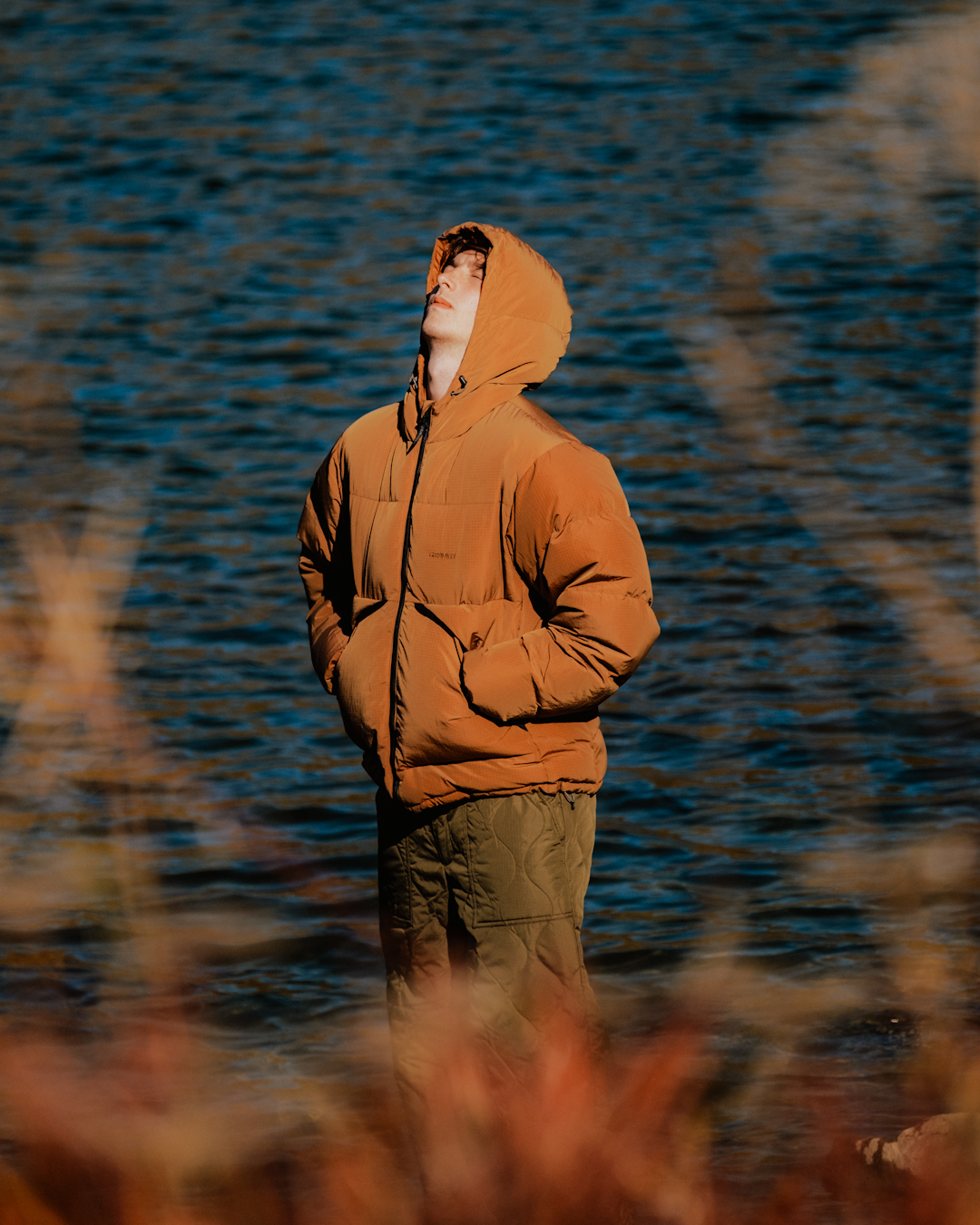 A man in a brown hooded puffer jacket and olive quilted pants standing by water in sunlight.