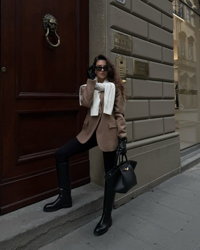 Woman in a camel blazer and black leggings with knee-high boots and a chunky scarf.
