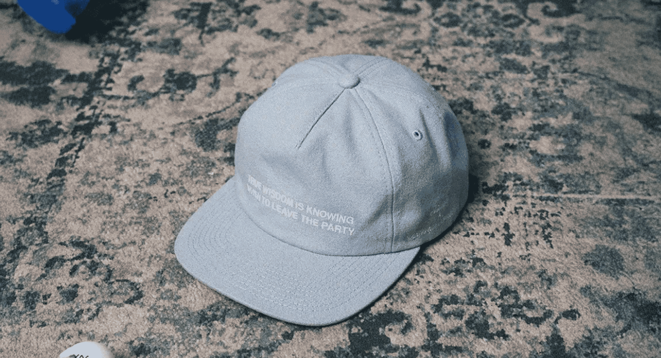Gray Stiksen baseball cap with white embroidered text reading 'TRUE WISDOM IS KNOWING WHEN TO LEAVE THE PARTY' on a patterned carpet.