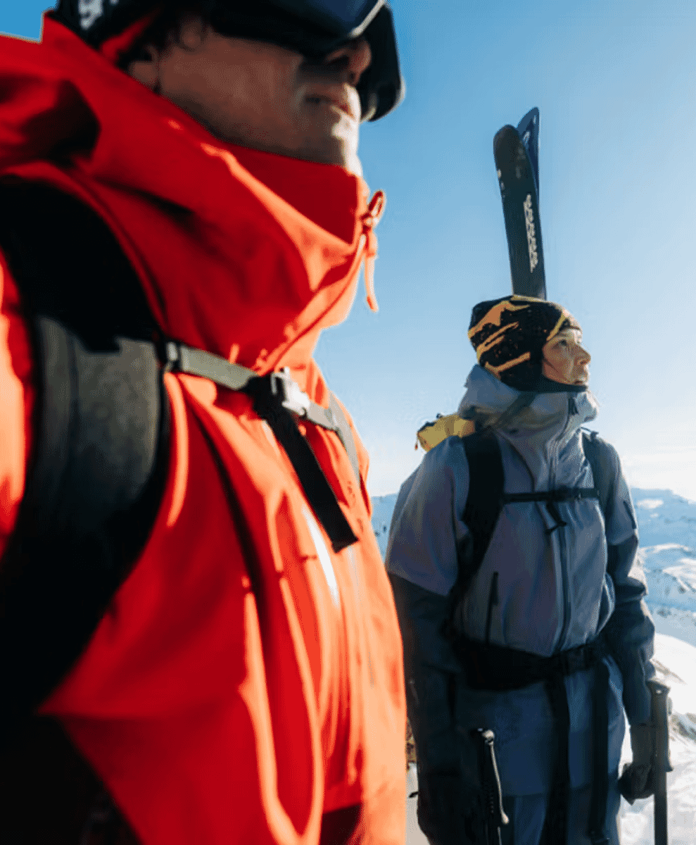 Two skiers in high-performance gear stand on a snowy mountain under clear blue skies.