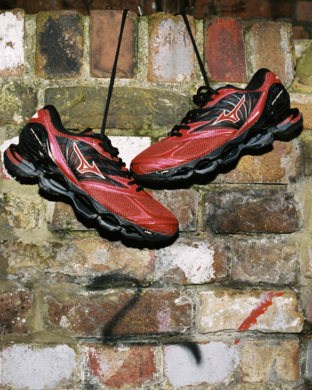Red and black Mizuno sneakers hanging by laces against an aged brick wall.