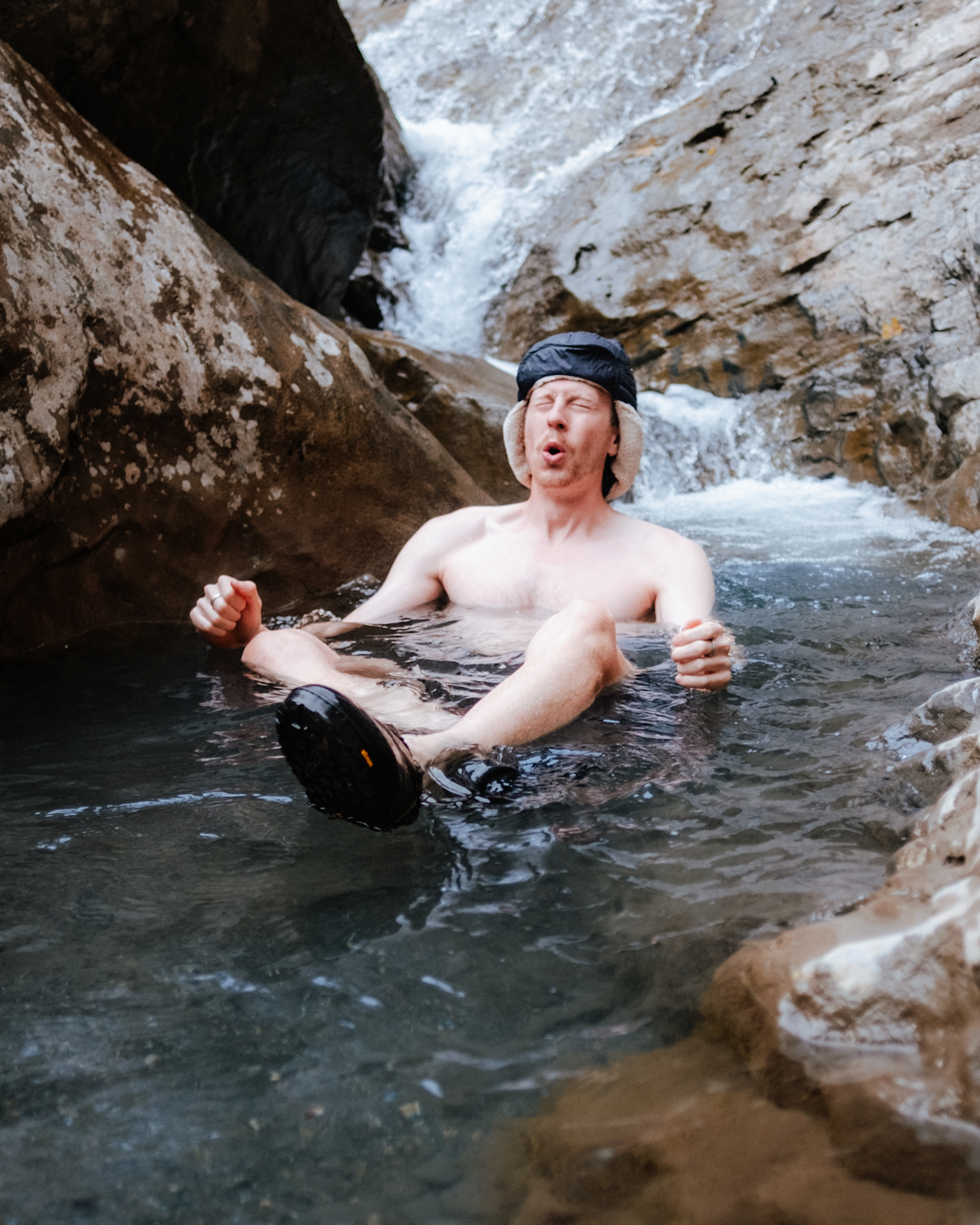 A man wearing a black fur-lined hat and hiking shoes sitting in a cold mountain stream.