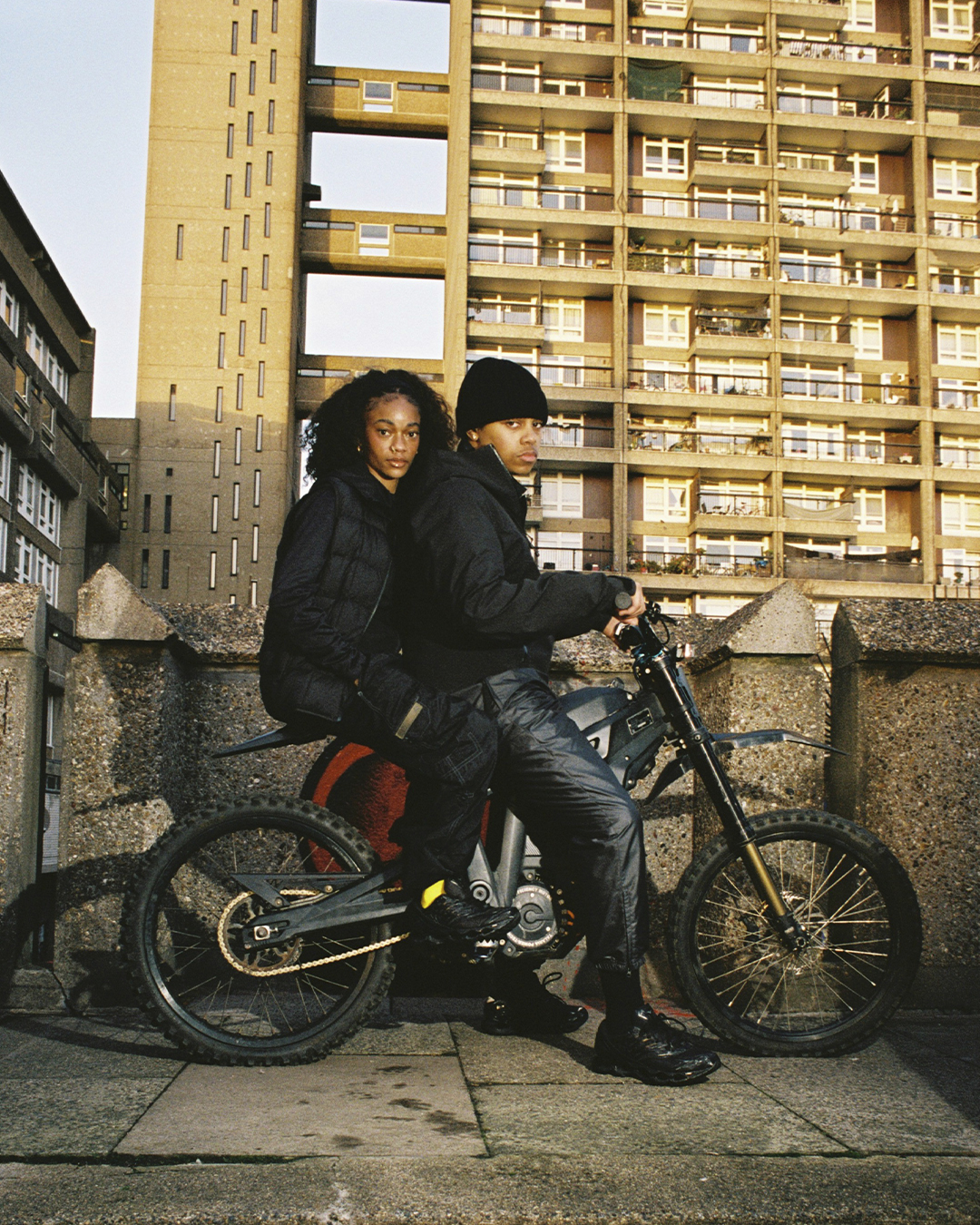 Two people in dark technical outerwear posing on a motorcycle in front of a Brutalist concrete building.